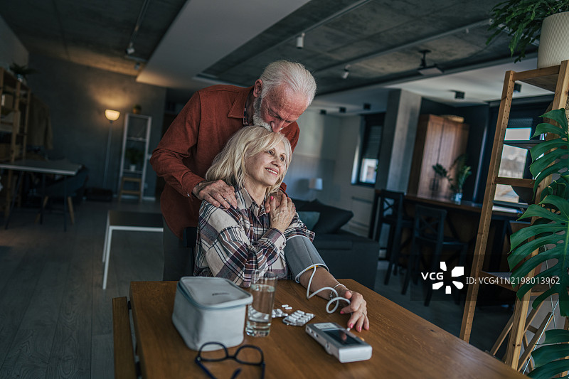 一位年长女性坐在桌子旁，使用数字血压监测仪测量自己的血压。她的丈夫站在她身后，轻轻地吻着她的头。图片素材