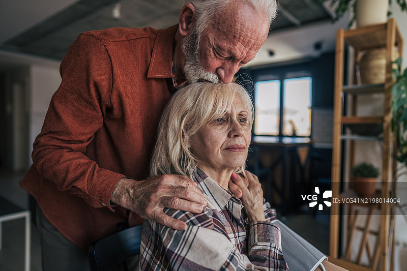 一位年长女性坐在桌子旁，使用数字血压监测仪测量自己的血压。她的丈夫站在她身后，轻轻亲吻她的头。图片素材