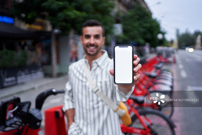 年轻人在城市环境中微笑着看向镜头，手持智能手机对着相机。图片素材