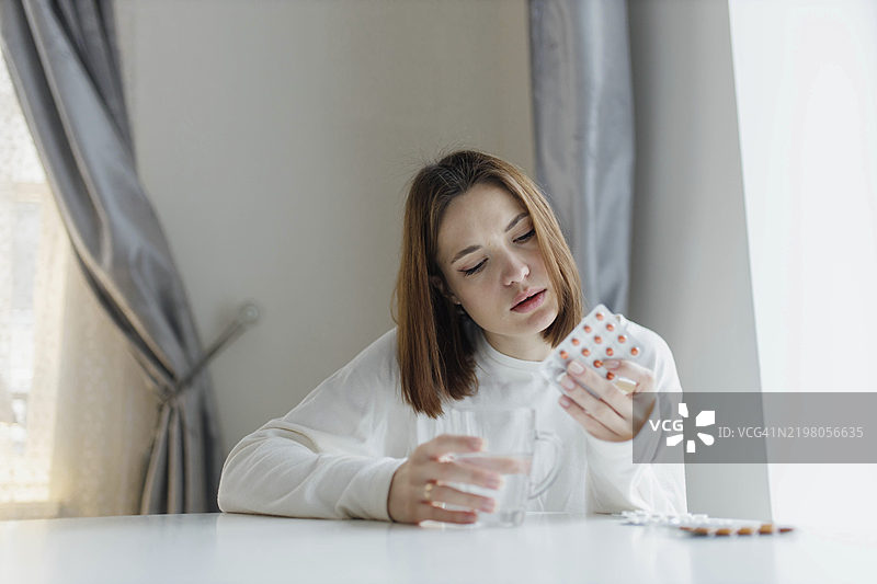 女人坐在桌子旁，手中检查一包药片，旁边有一杯水。图片素材