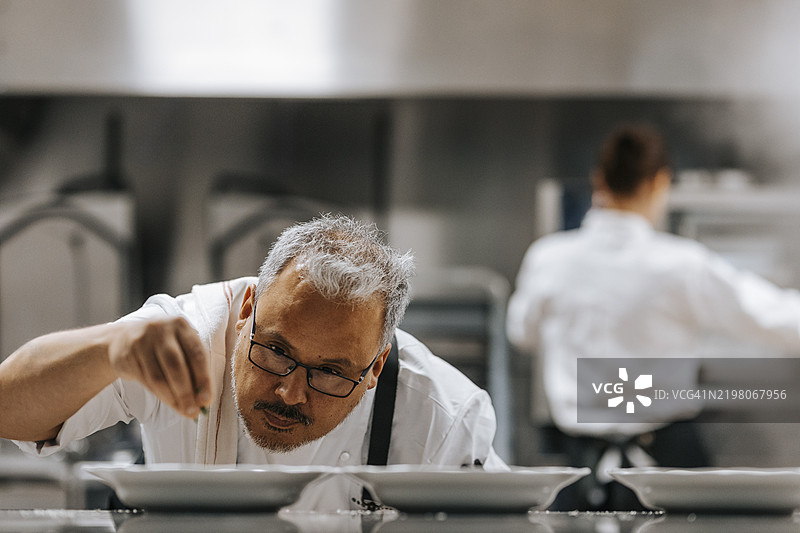 成熟男性厨师戴着眼镜，在商业厨房中为食物装饰。图片素材