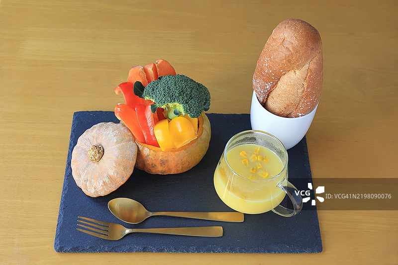 静物：西兰花、玉米浓汤、法棍、整只南瓜、黑盘上的红椒粉，金色叉子和勺子，棕色背景图片素材