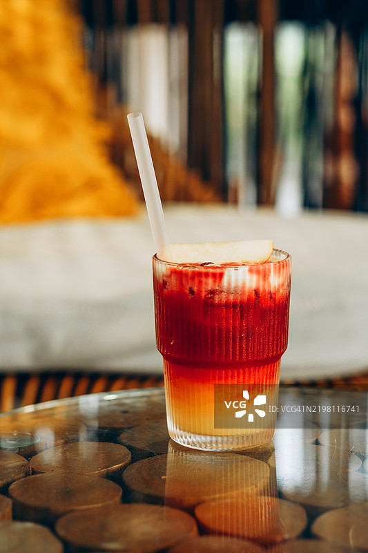 玻璃杯中分层的冰饮，红色和黄色渐变，放在玻璃桌上。可见塑料吸管和冰块。室内环境，背景模糊的靠垫和木质装饰。图片素材