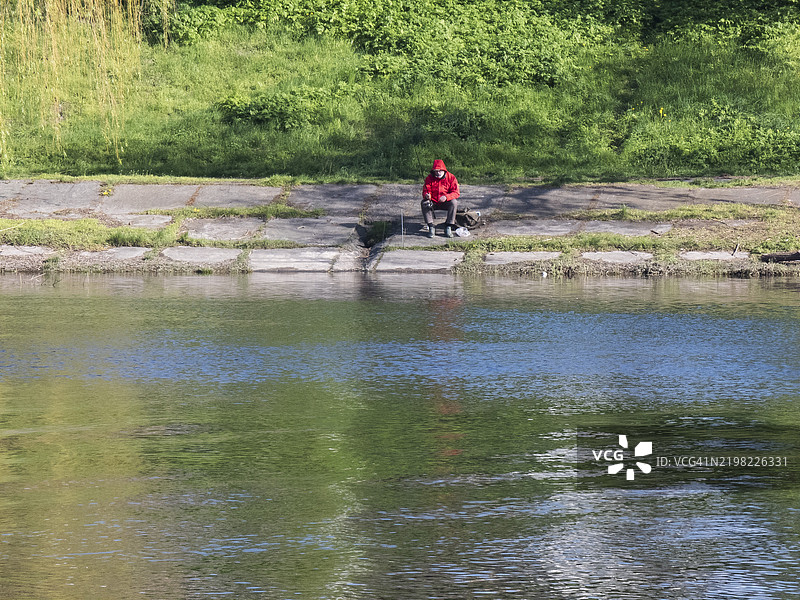 渔夫穿着鲜红色夹克，静静地坐在河岸上，周围是大自然的美景，正值正午。图片素材