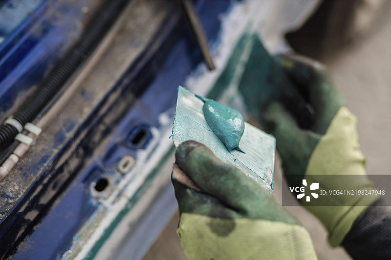 戴手套的手在车上涂抹青色填料，周围有工具，工作室环境。图片素材