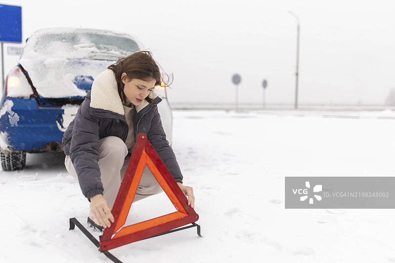 一名女性在雪-covered的道路上放置警告三角架，附近停着一辆车。图片素材