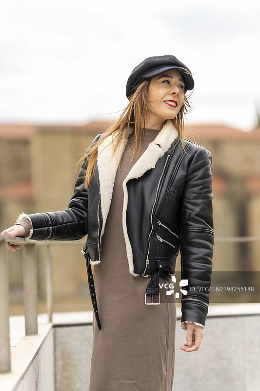 时尚女性在城市环境中摆姿势，穿着黑色皮夹克，搭配羊毛领和报童帽。图片素材