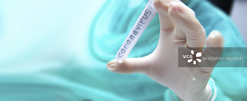 戴手套的手握着冠状病毒试管，背景中有一个穿着蓝绿色服装的人。图片素材