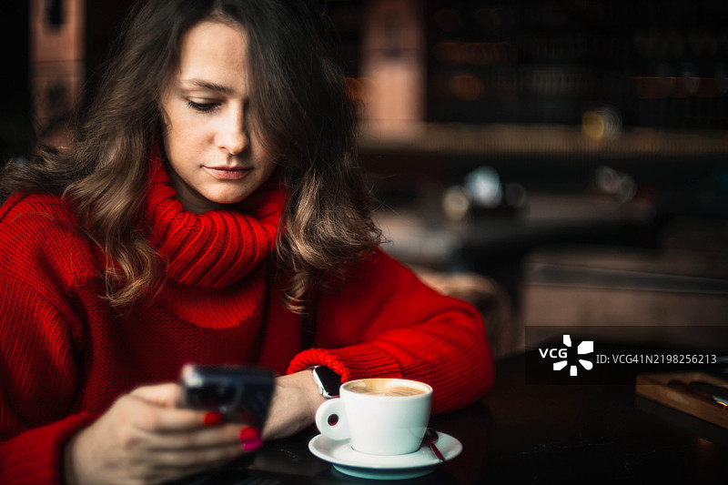 穿着红色毛衣的女性在咖啡馆专注于她的智能手机，旁边有咖啡。图片素材