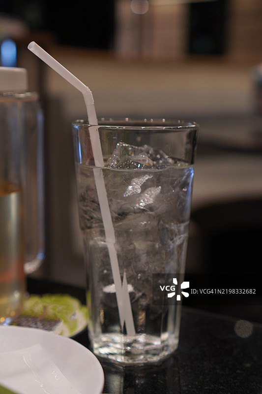 清澈玻璃杯中装有冰块的清凉饮料，配有塑料吸管，放在木桌上。图片素材