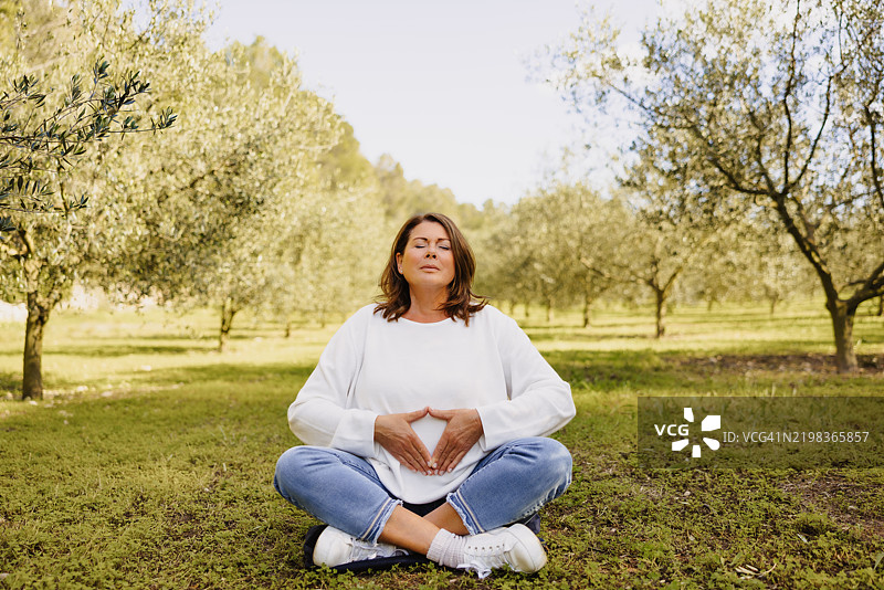 成熟的女性在更年期，盘腿坐在莲花坐姿中，双手形成神圣的三角手印，周围是年轻的橄榄树和生机勃勃的绿意。图片素材