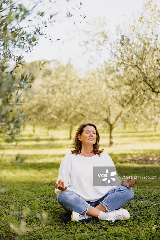 一位成熟的女性在更年期，坐在莲花坐姿中，双手做着智慧印，周围环绕着柔和的年轻橄榄树绿意，享受宁静的时刻。图片素材