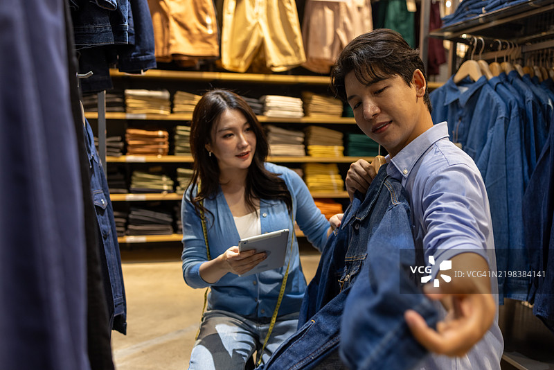 亚洲女性顾客在商店选择服装尺码，亚洲男性销售员推荐产品并查看数字平板上的服装样本。图片素材