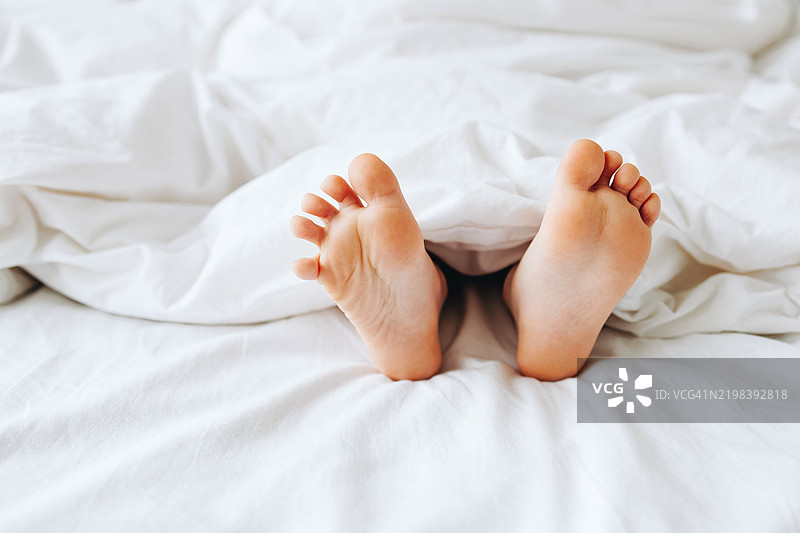 小脚丫从温暖的白色毯子下露出来，体现了童年、家庭舒适和宁静休息的概念。图片素材