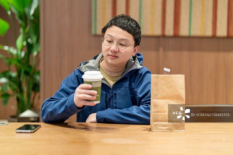 一位亚洲男子在咖啡店里，手里拿着咖啡，旁边有一个纸袋和手机。图片素材