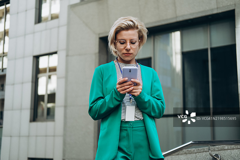 身穿青绿色西装的年轻女性在商务中心前使用智能手机。图片素材
