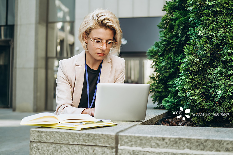 年轻女性穿着浅色西装外套，在商业中心附近的柏树旁使用超极本工作。图片素材