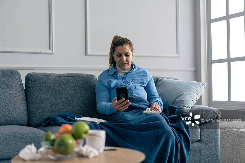 女人坐在沙发上，神情疲惫，手里拿着手机和纸巾，周围是温馨的家居环境图片素材