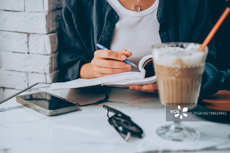 年轻女性在咖啡馆桌子上写书，旁边有手机、冰咖啡和太阳镜。图片素材