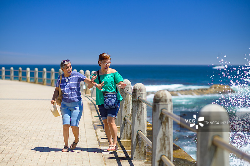 两位年长女性在海浪冲击和溅起水花时举起双臂图片素材