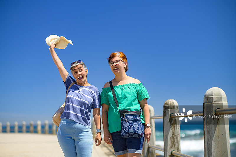 两位年长女性在海滨长廊上，手臂抬起，戴着帽子。图片素材