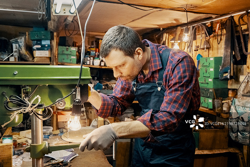 精密钻孔在地下室工作区进行。中年修理工穿着工装，专注于细致的机器操作。图片素材