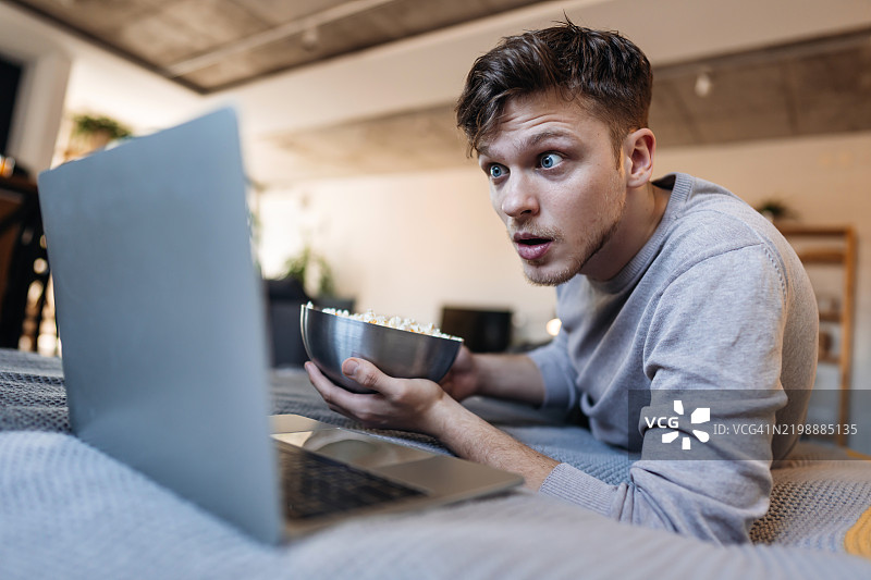 惊讶的年轻男子在笔记本电脑上观看电影并吃着爆米花图片素材