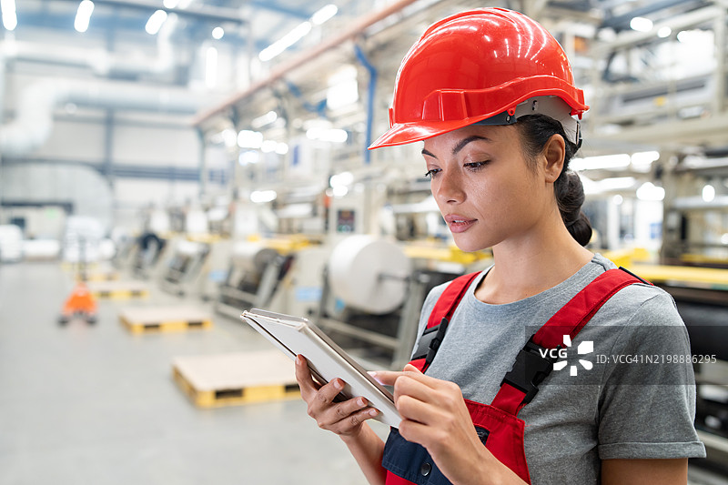 穿着红色防护装备的女性工人在大型工厂中使用平板电脑，专注而精准。图片素材