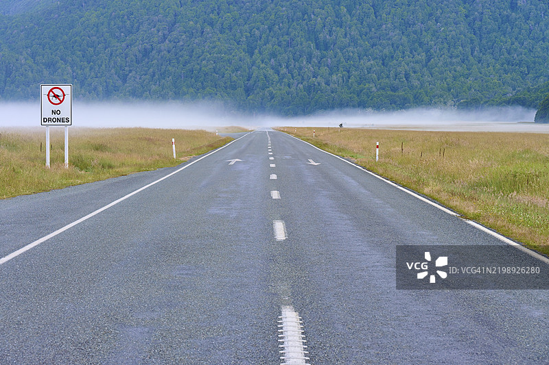 雾气弥漫的风景中,笔直的沥青道路旁有无人机禁飞标志,夏季,埃格林顿谷观景点,米尔福德峡湾,提阿瑙山下,菲ordland国家公园,新西兰南岛,南太平洋。图片素材