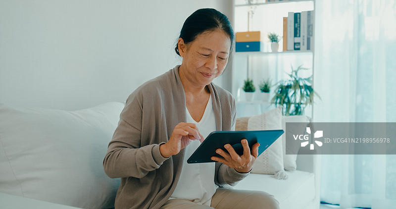 亚洲微笑的老年女性在家中的沙发上使用平板电脑，科技在提升老年人福祉和连接性方面的作用。图片素材