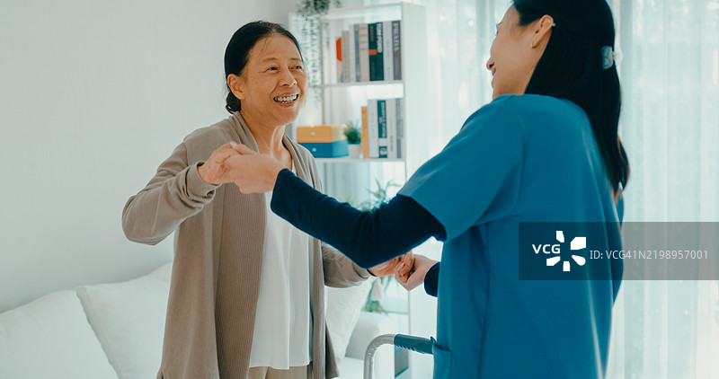 年轻的亚洲护士和一位老年女性在家中的客厅里共享快乐的舞蹈时光，展现了陪伴和积极的生活方式。家庭健康护理。图片素材