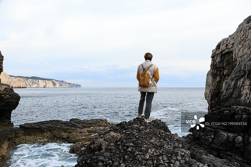 一位成熟的女性游客站在黑色岩石上，背对着海洋。图片素材
