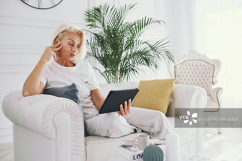 年长女性坐在舒适的沙发上放松，使用平板电脑与居住在国外的家人进行视频通话，享受远程交流的乐趣。图片素材