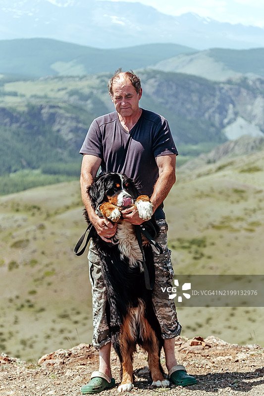中年男子顽皮地将他的伯恩山犬举起，背景是令人惊叹的绿色山谷和远处的丘陵。图片素材