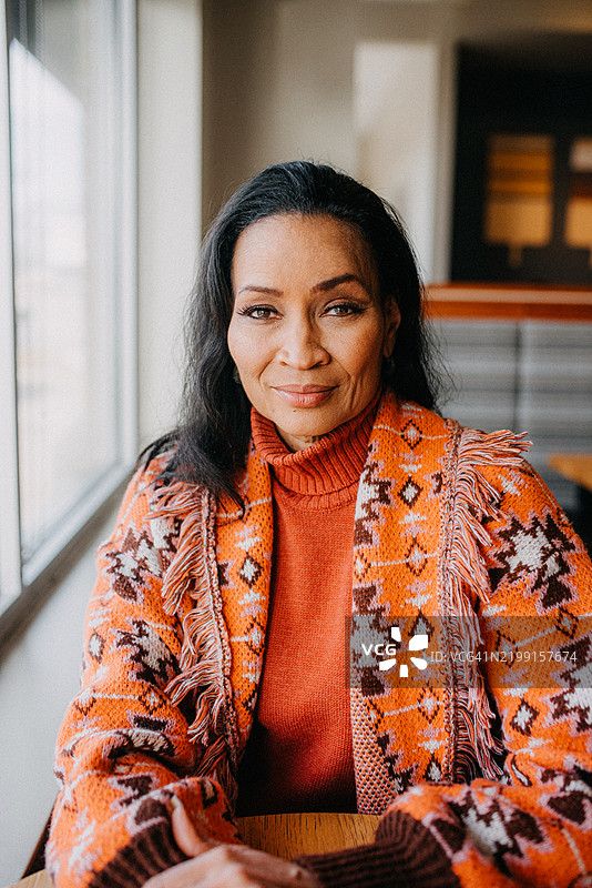 一位穿着橙色图案毛衣的女性自信地坐在靠窗的桌子旁，自然光线勾勒出她的面容。图片素材