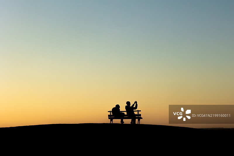 夕阳下，情侣坐在长椅上，剪影映衬着天空图片素材