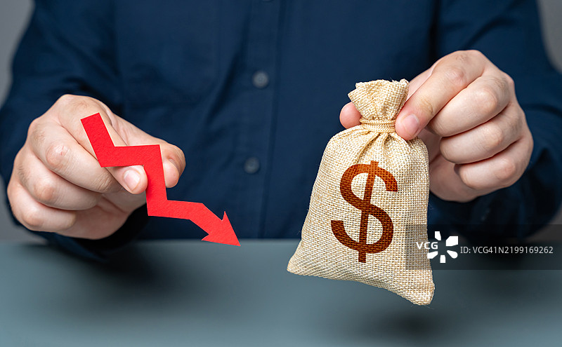 一名男子手持一支指向下方的红色箭头，旁边有一个美元钱袋，象征着财务困难、损失和盈利能力下降，经济衰退、薪资削减、财务压力和货币贬值。图片素材