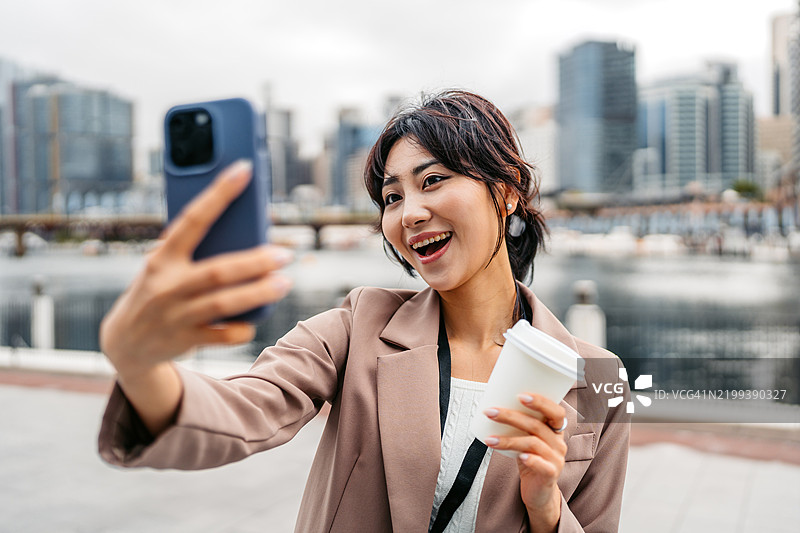 年轻的亚洲女商人在澳大利亚悉尼达令港一边喝外带咖啡一边用手机自拍图片素材