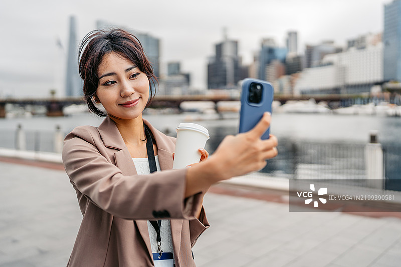 年轻的亚洲女商人在澳大利亚悉尼达令港一边喝外带咖啡一边用手机自拍图片素材