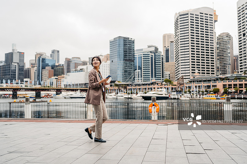 年轻的亚洲女商人在澳大利亚悉尼达令港边走边喝咖啡并使用手机图片素材
