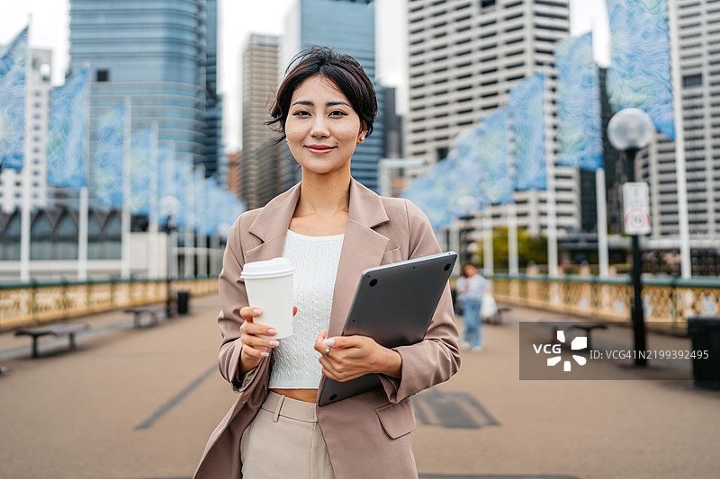 年轻的亚洲女商人在澳大利亚悉尼达令港一边喝外带咖啡，一边拿着笔记本电脑。图片素材