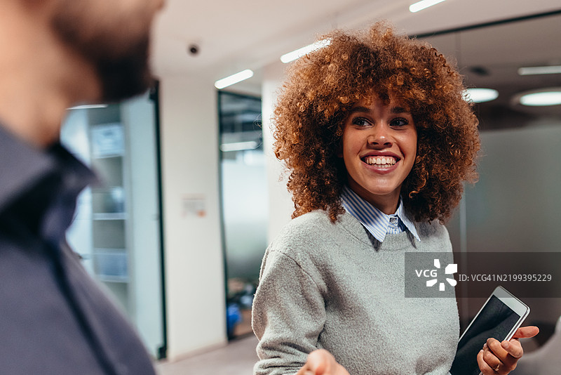 年轻女商人手持平板，微笑着看着现代办公室里的同事图片素材