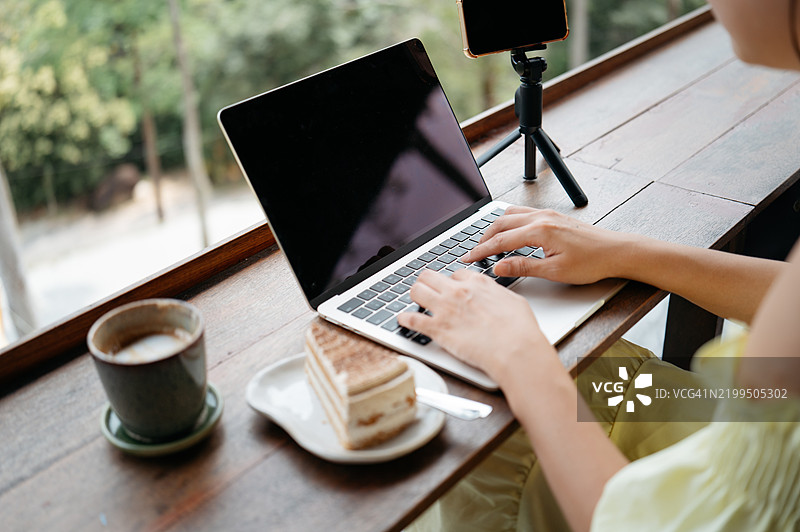 亚洲女性旅行者在咖啡馆阳台露台上休息喝茶，同时使用笔记本电脑。图片素材