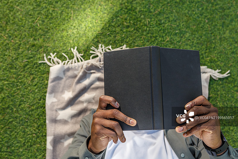 一个人坐在柔软的毯子上，沉浸在郁郁葱葱的绿草地上，专心阅读一本书。图片素材