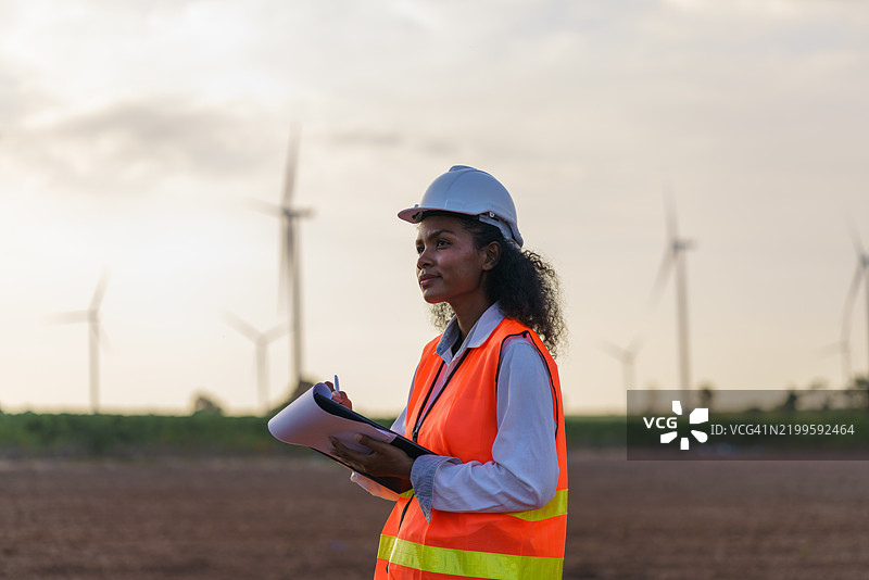 一位年轻的非裔美国工程师正在记录风电场调查的数据，背景是夕阳下的风力涡轮发电业务。图片素材