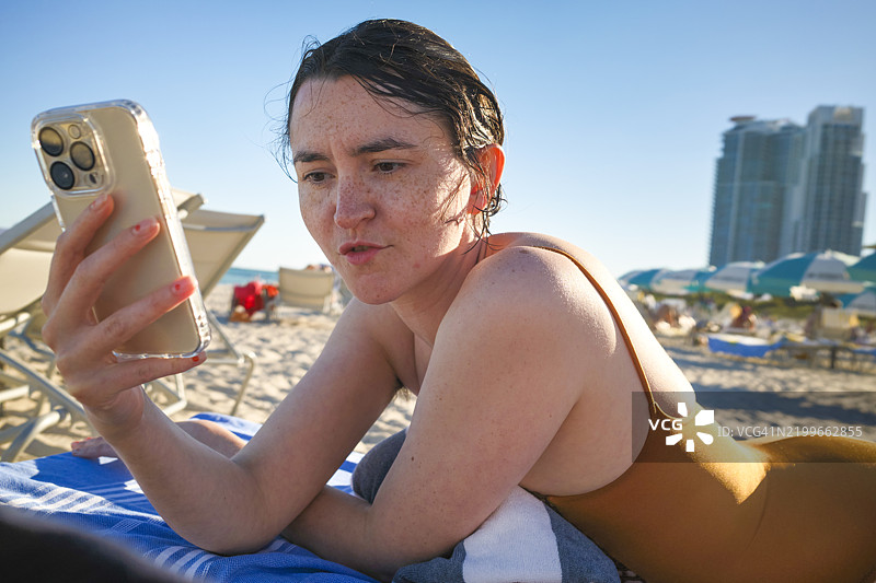 穿着泳衣的女人躺在海滩上，面带独特表情地看着智能手机，背景是阳光明媚的天空，佛罗里达州迈阿密。图片素材