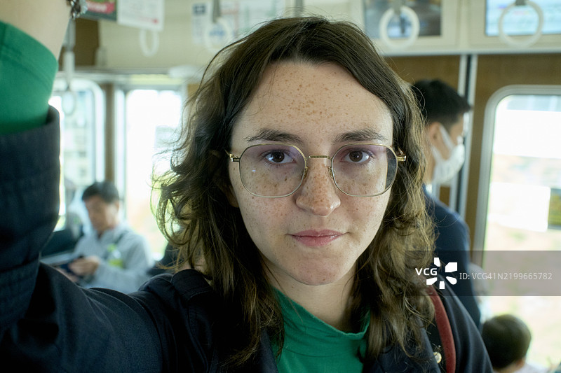 戴眼镜的女性在拥挤的火车上握着扶手，直视镜头。日本大阪图片素材