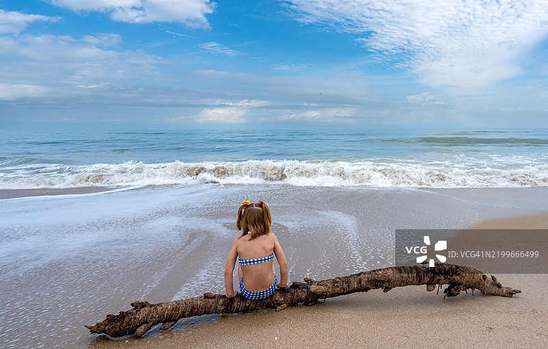 年轻女孩坐在佛罗里达州梅里特岛卡纳维拉尔国家海岸的海滩上漂流木上，清晨的亚特兰大海洋旁。图片素材