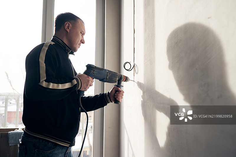 一个男人正在用电锤打墙，并为灯具铺设电缆。图片素材