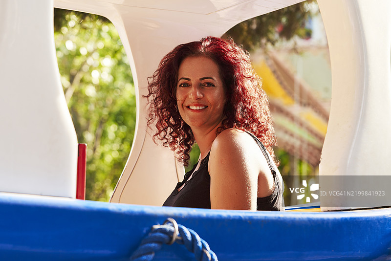 一位穿着休闲服的美丽女性坐在游乐园儿童列车上，微笑着侧身看向镜头。图片素材